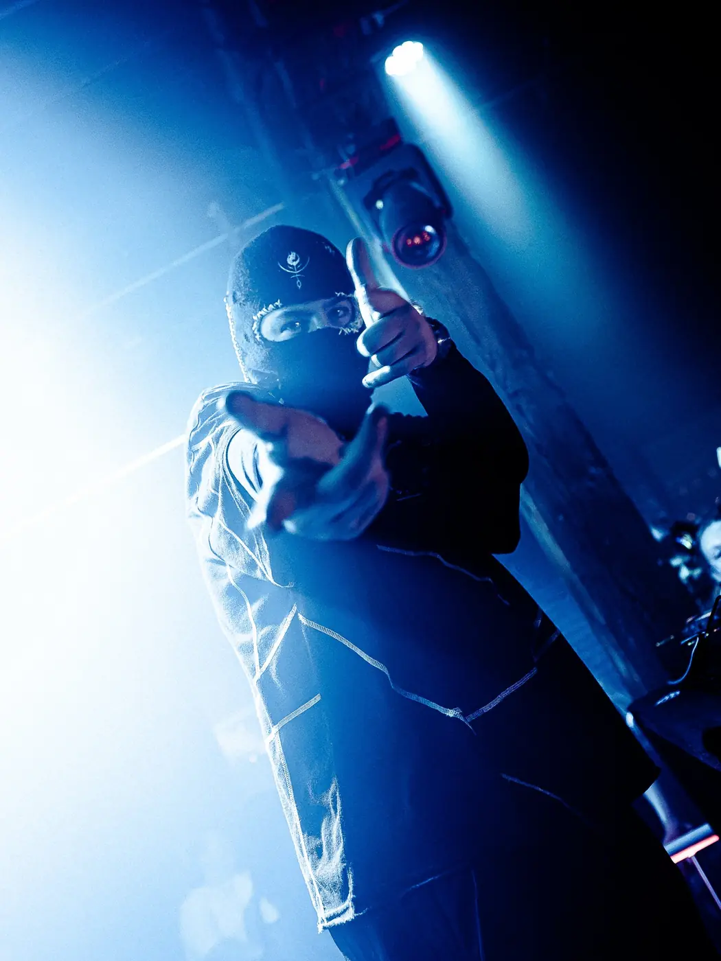 Masked performer pointing at the camera under blue light